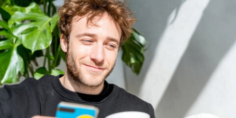 Sunlit room, European male joyfully checks vibrant emoji phone, embodying Digital Detox Day relaxation and quirky tech culture