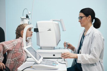 Caucasian female child undergoing eye examination with ophthalmologist using advanced diagnostic equipment in medical clinic, doctor operating machine and monitoring results
