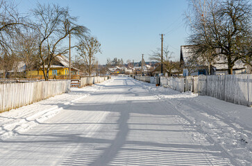 A snow-covered village street lined with wooden fences and bare trees under a clear winter sky,...