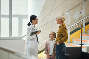 Caucasian middle aged woman and Caucasian child girl standing and talking with young female doctor holding digital tablet in modern medical facility near staircase