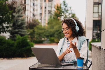 Young woman with headphones working on a laptop outdoors at a modern city cafe enjoying a sunny...