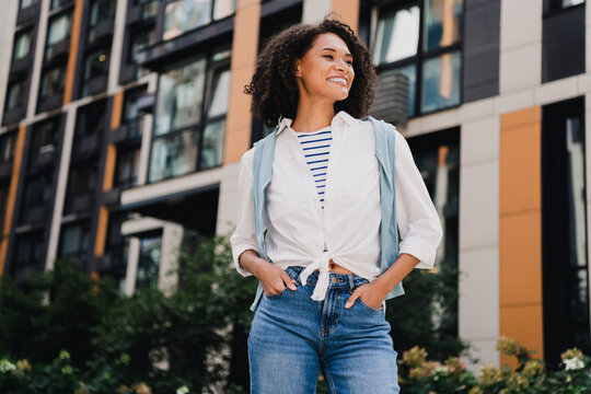 Young woman with backpack in urban city setting smiles while walking in casual fashion beside modern buildings and colorful architecture in a vibrant street scene