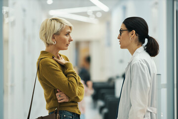 Caucasian middle aged woman talking to young adult Asian female doctor in medical facility hallway, both standing and facing each other, discussing health related concerns