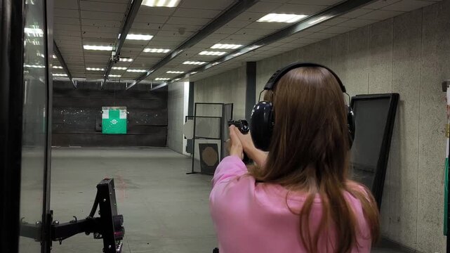 A woman in protective headphones shoots a pistol at a target in a closed shooting range. Training and safety, view from the back, the flash of a shot is noticeable.
