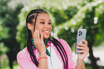 Smiling young woman with braided hair using a smartphone in a sunny park showcasing casual fashion outdoor lifestyle and modern urban vibe