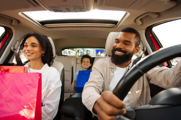 Family shopping day. Happy Latin parents and son riding car holding colorful shopper bags, sitting in vehicle and smiling