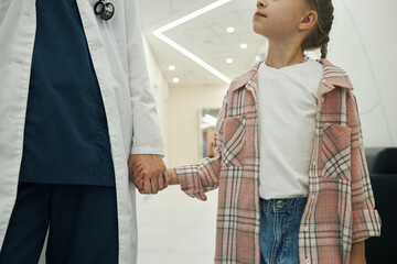 Caucasian female doctor holding hand of Caucasian child girl while walking together in hospital...