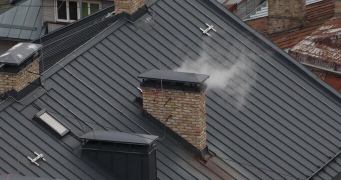 Close view of white smoke venting from a brick chimney on a dark metal rooftop. Cold, overcast light suggests winter heating, urban routine, and energy use across old buildings.