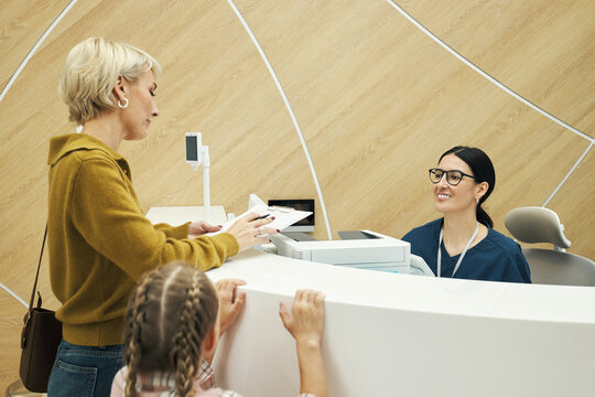 Caucasian middle aged woman handing documents to smiling Caucasian young adult woman receptionist, while Caucasian girl standing nearby, at front desk in modern medical office