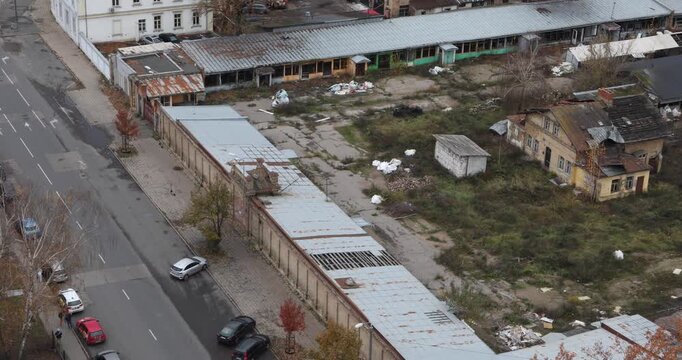 High-angle view of an abandoned industrial yard with rusted roofs, broken sheds and scattered trash amid puddles and weeds. Overcast light suggests neglect, hardship and slow transition.