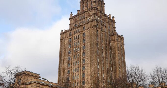 Close-up of the Latvian Academy of Sciences, a Soviet-era Stalinist tower rising against bright clouds in Riga. Ornate tiers and a sharp spire show monumental socialist classicism and layered history.