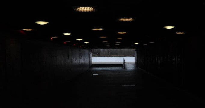 View from inside a dark pedestrian underpass toward a river; rows of ceiling lights lead to a bright exit. The moody contrast and empty walkway suggest solitude, transition, and a pull toward light.