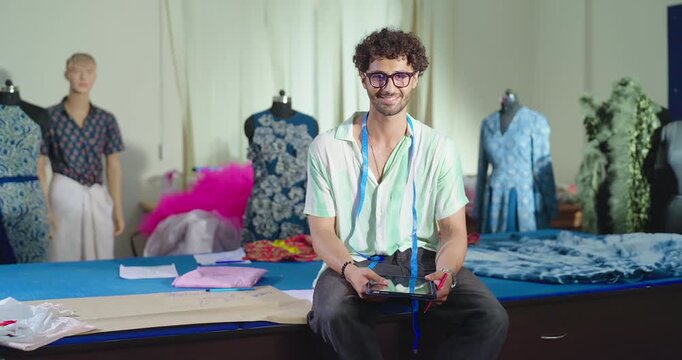 Indian young adult gen z smart male man hold tab learn new clothe craft course look camera class room. Boy sitting on desk table wear glass tape around neck staring cam do causal pose enjoy day place