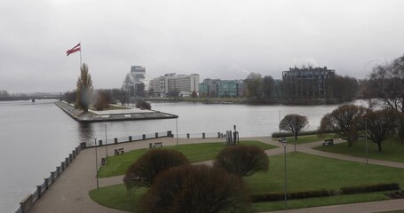 Riga’s riverside park with trimmed lawns, winding paths and a large Latvian flag near the National Library of Latvia. Calm water and soft overcast light evoke civic pride and quiet autumn weather.