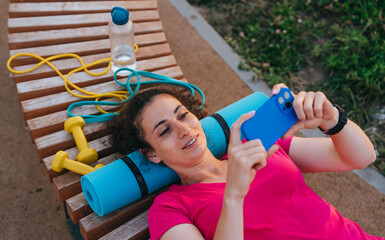 Woman lies on a bench, surrounded by fitness gear, comfortably engaging with her phone during a...