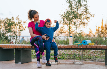 Mother and child on a bench with fitness gear, joyfully flexing muscles and sharing laughter in a serene outdoor setting.