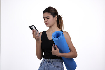 Young woman holding a blue yoga mat and looking at her smartphone with a confused or frustrated...
