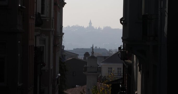 Between two old facades, birds glides through a hazy street canyon as a hilltop tower fades in distance. Soft backlight, layered rooftops, and morning mist suggest quiet travel, pause, and longing.