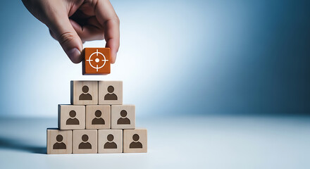 Hand placing a wooden block with a target icon on top of a pyramid of blocks with person icons, symbolizing team goals and leadership.