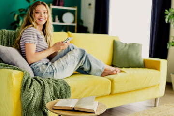 Relaxed woman at home lounging on a bright yellow sofa with a book and phone in a cozy living room setting