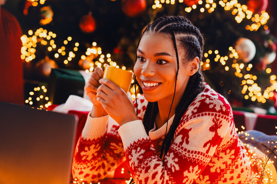 Cheerful woman at home enjoys festive season smiles while sipping from a mug beside a laptop near a warmly lit Christmas tree