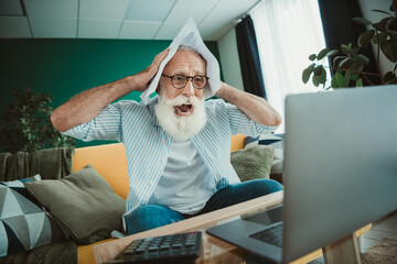 Surprised senior man at home working on laptop with paper on head in cozy living room during a casual day