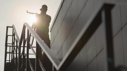 Silhouette of Black young man carrying bicycle going down stairs of grey office building against shining sun on summer day - Powered by Adobe