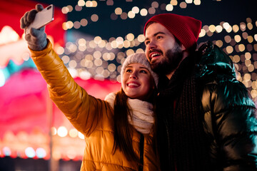 Funny couple selfie at festive market night with Christmas lights and snowy coats capturing love...