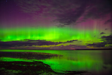 Vivid northern lights glowing green and purple over a calm lake with reflections and scattered...