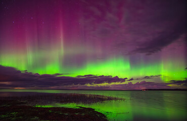 Brilliant aurora borealis in green and purple hues shines above a calm lake and cloudy night sky...