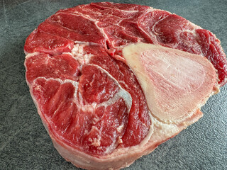Raw beef shank cross-cut with bone on dark stone countertop, high-angle side view showing marbled meat texture for culinary use and food stock photography