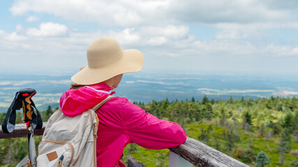 Rear view of a woman hiker in a wide-brim hat and pink jacket resting at a viewpoint, backpack and...