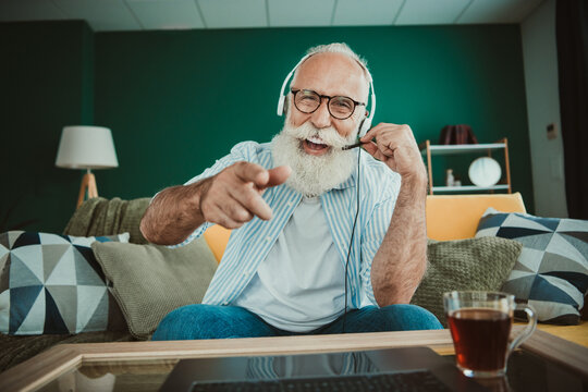 Joyful senior man with headset plays video game at home in a cozy living room smiling and pointing at the camera - Powered by Adobe