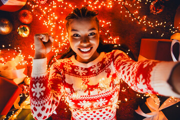 Cheerful woman in red white christmas sweater takes a selfie at home surrounded by fairy lights gifts and warm holiday decor
