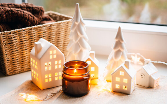 A warm holiday vignette featuring ceramic houses, soft candlelight, and decorative trees arranged by a windowsill. Perfect for festive decor, homey atmosphere, and Christmas-themed marketing materials