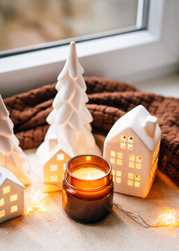 A warm holiday vignette featuring ceramic houses, soft candlelight, and decorative trees arranged by a windowsill. Perfect for festive decor, homey atmosphere, and Christmas-themed marketing materials