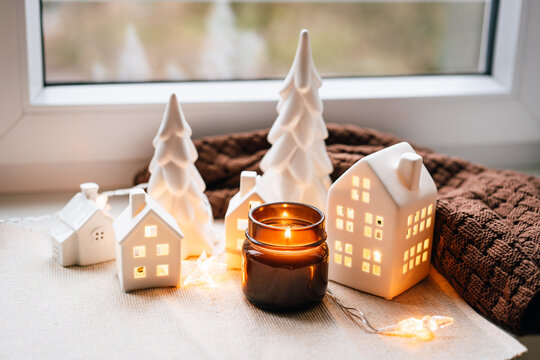 A warm holiday vignette featuring ceramic houses, soft candlelight, and decorative trees arranged by a windowsill. Perfect for festive decor, homey atmosphere, and Christmas-themed marketing materials