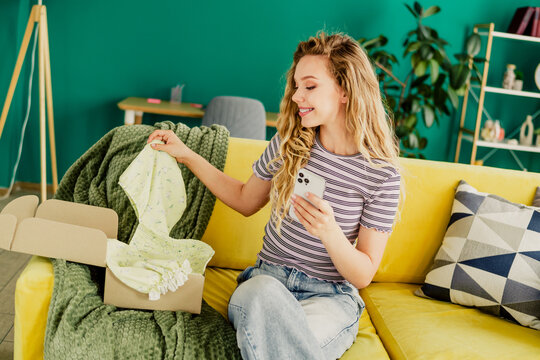 Bright modern living room scene with a cheerful young woman on a yellow couch checking her phone while unboxing a cozy green blanket