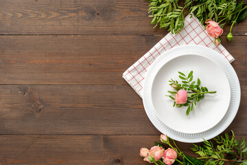 Top view of table setting with empty plate and napkin - pink roses flowers decorated on brown wooden background, flat lay