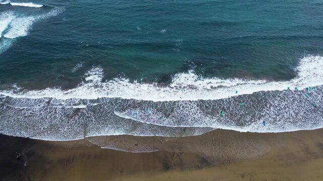 overhead aerial (top-down) view of the surfing section of a sandy beach (likely La C&iacute;cer, Las Canteras). The image cleanly separates the dark brown sand, the white foam of the breaking waves,