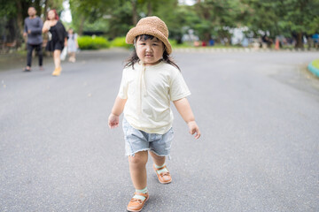 Fototapeta premium A cute little Asian girl wearing a straw hat walks happily on a walkway in a park.