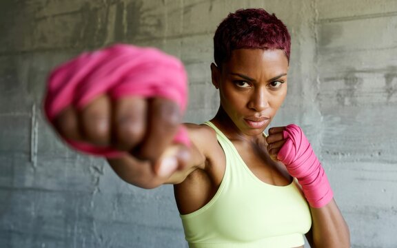 Determined woman punching with pink hand wraps against a concrete wall