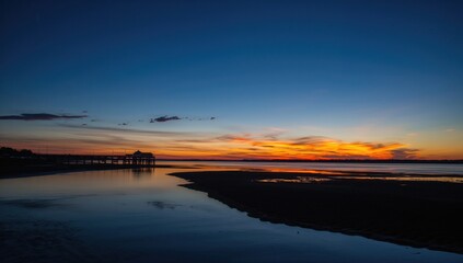 Tranquil river shore at dusk featuring a darkened pier, still water mirroring the warm colors of the evening sky, and a serene sandy area.