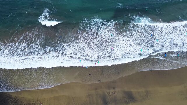 overhead aerial (top-down) view of the active surfing area at a sandy beach (La C&iacute;cer, Las Canteras). The composition is horizontally divided into the dark wet sand, the foamy white line of the waves