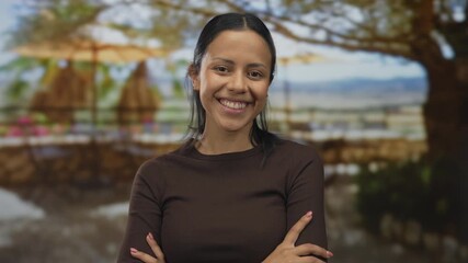 Young hispanic woman smiling confidently outdoors with arms crossed under a tree on a sunny day where colorful flowers bloom elegantly in the blurred background. - Powered by Adobe