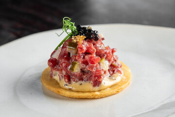 gourmet tartare dish, garnished with black caviar and microgreens, served on a crisp cracker on a white plate with a dark background