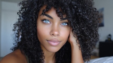 Fototapeta premium A woman with voluminous curly hair rests her hand on her face, showcasing beautiful blue eyes and a warm smile. This scene captures a relaxed moment in a bright indoor space.