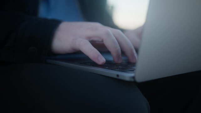 Close-up side angle of a person's hands typing on a slim laptop while sitting outdoors, suggesting remote work or study.  

