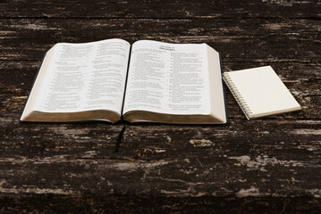 Flat lay of open Holy Bible (Psalms 149-150, Proverbs 1) and notepad on rustic wood table. Top view in natural daylight. Symbolizes spiritual study, wisdom, reflection, and devotion.
