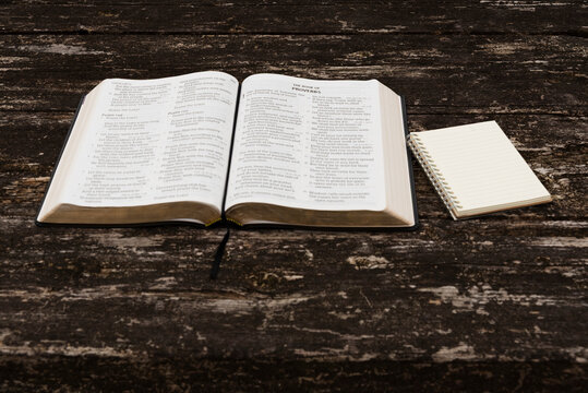 Flat lay of open Holy Bible (Psalms 149-150, Proverbs 1) and notepad on rustic wood table. Top view in natural daylight. Symbolizes spiritual study, wisdom, reflection, and devotion.